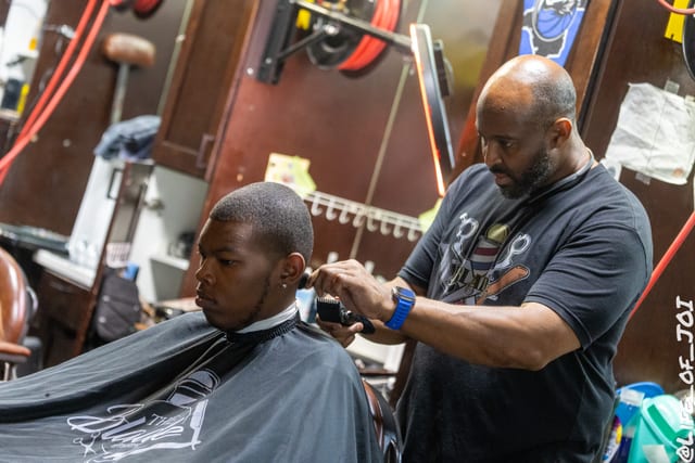 A fade takes shape as a barber works on a student wearing a Blade Barbershop cape.